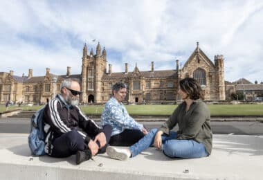 Three students sitting and laughing in front of an old building on University of Sydney campus.