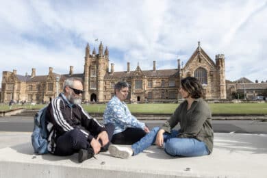 Three students sitting and laughing in front of an old building on University of Sydney campus.