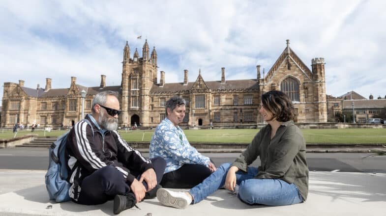 Three students sitting and laughing in front of an old building on University of Sydney campus.