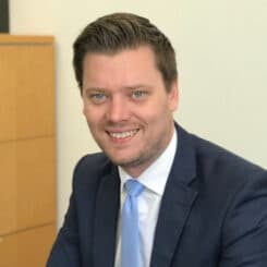 A picture of Jarrod smiling, he is wearing a suit with a blue tie